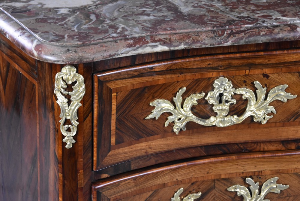 An 18th-century French Regence serpentine commode crafted in rosewood parquetry with gilded ormolu drawer pulls and a contoured marble top. View IX