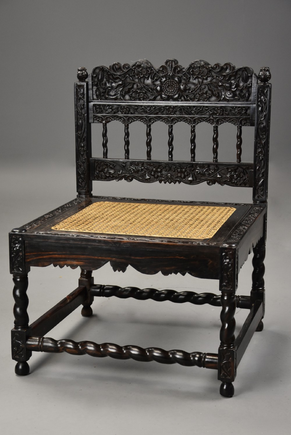 A 17th-century dark carved ebony low chair featuring floral relief carving on the frame, a hand-woven cane seat, and spiralled barley-twist legs.