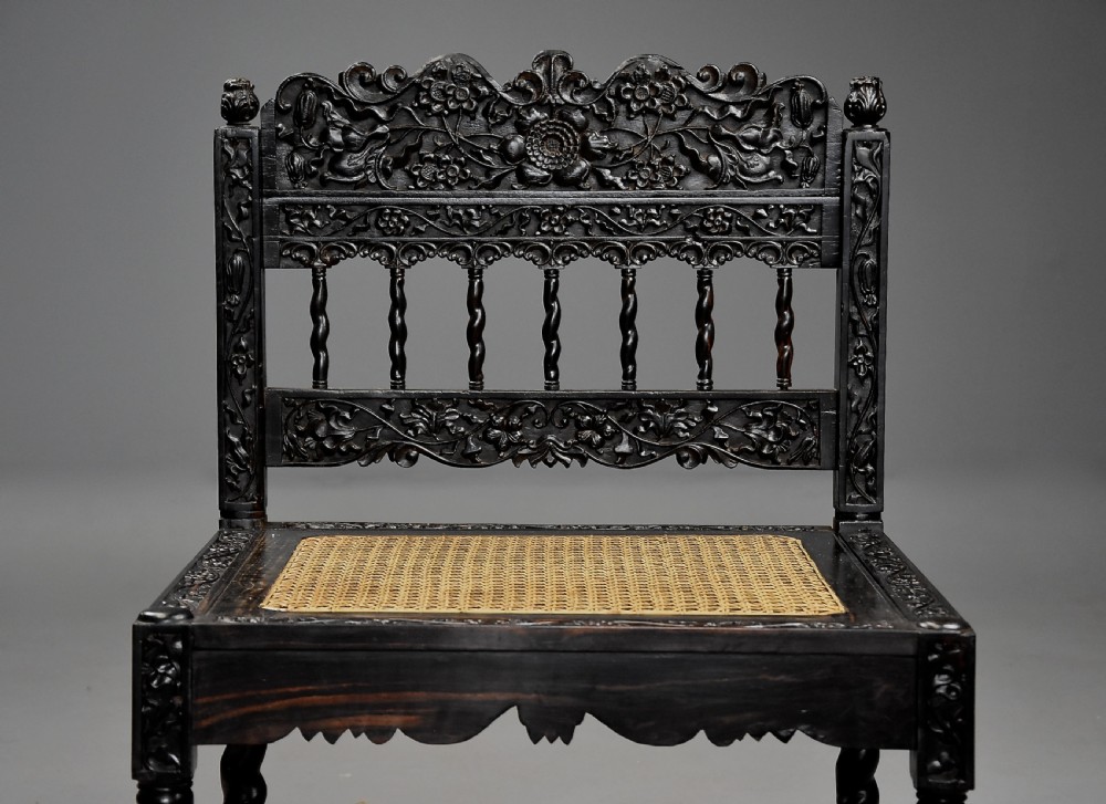 A 17th-century dark carved ebony low chair featuring floral relief carving on the frame, a hand-woven cane seat, and spiralled barley-twist legs. View IV