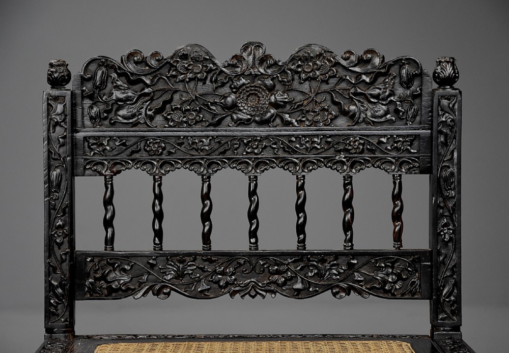 A 17th-century dark carved ebony low chair featuring floral relief carving on the frame, a hand-woven cane seat, and spiralled barley-twist legs. View V
