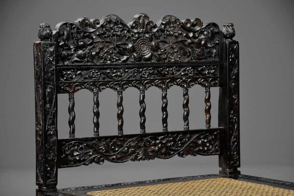 A 17th-century dark carved ebony low chair featuring floral relief carving on the frame, a hand-woven cane seat, and spiralled barley-twist legs. View VII