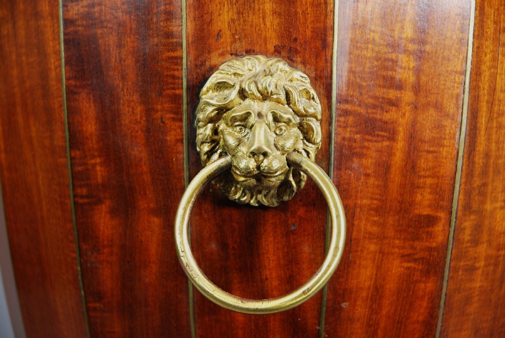 An 18th-century turned mahogany cutlery urn with brass stringing, lion mask ring handles, a top brass finial, and a stepped square base resting on brass ball feet. View VII