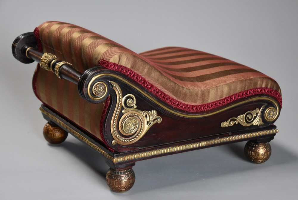A nineteenth-century Regency mahogany footstool featuring a scrolled top upholstered in red and gold striped fabric with ornate gilt-metal mounts and ball feet.