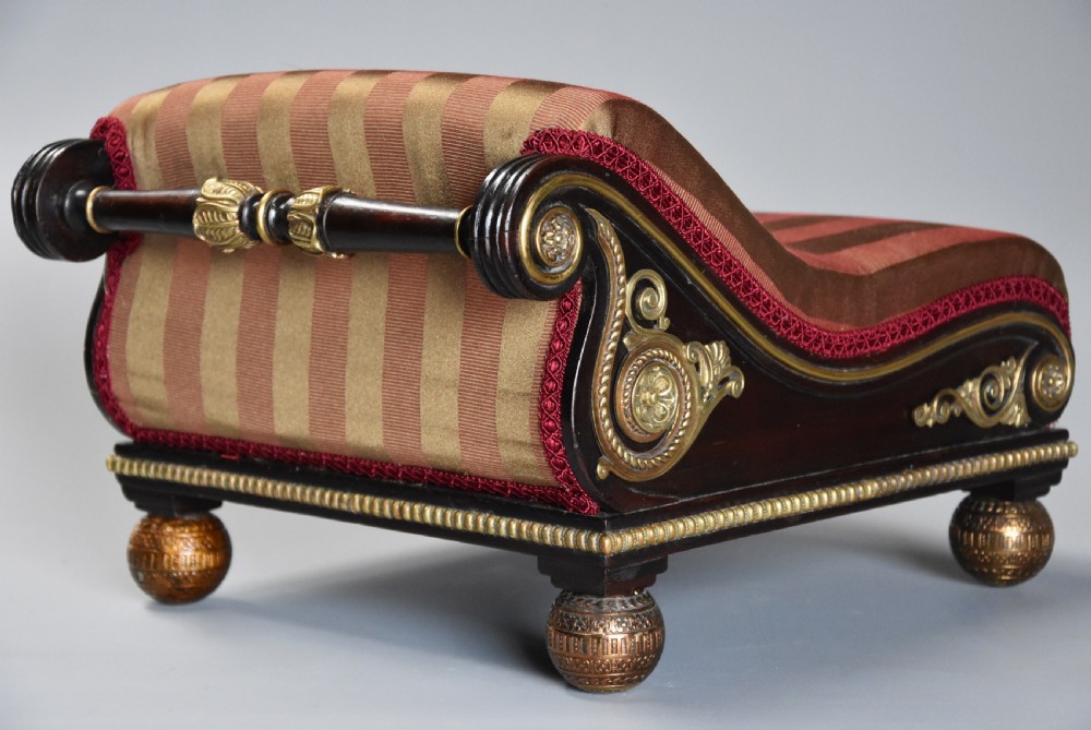 A nineteenth-century Regency mahogany footstool featuring a scrolled top upholstered in red and gold striped fabric with ornate gilt-metal mounts and ball feet. View II