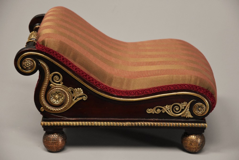 A nineteenth-century Regency mahogany footstool featuring a scrolled top upholstered in red and gold striped fabric with ornate gilt-metal mounts and ball feet. View VI