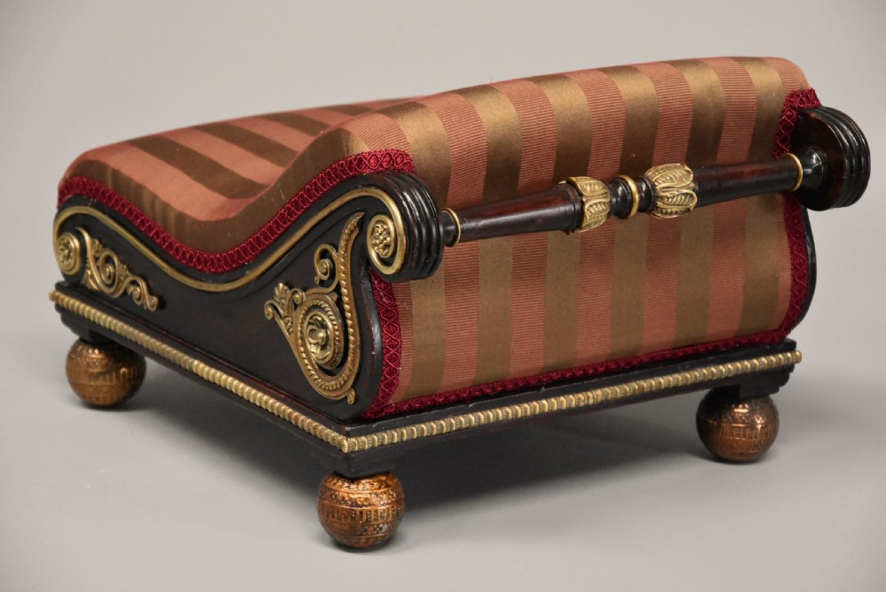 A nineteenth-century Regency mahogany footstool featuring a scrolled top upholstered in red and gold striped fabric with ornate gilt-metal mounts and ball feet. View X