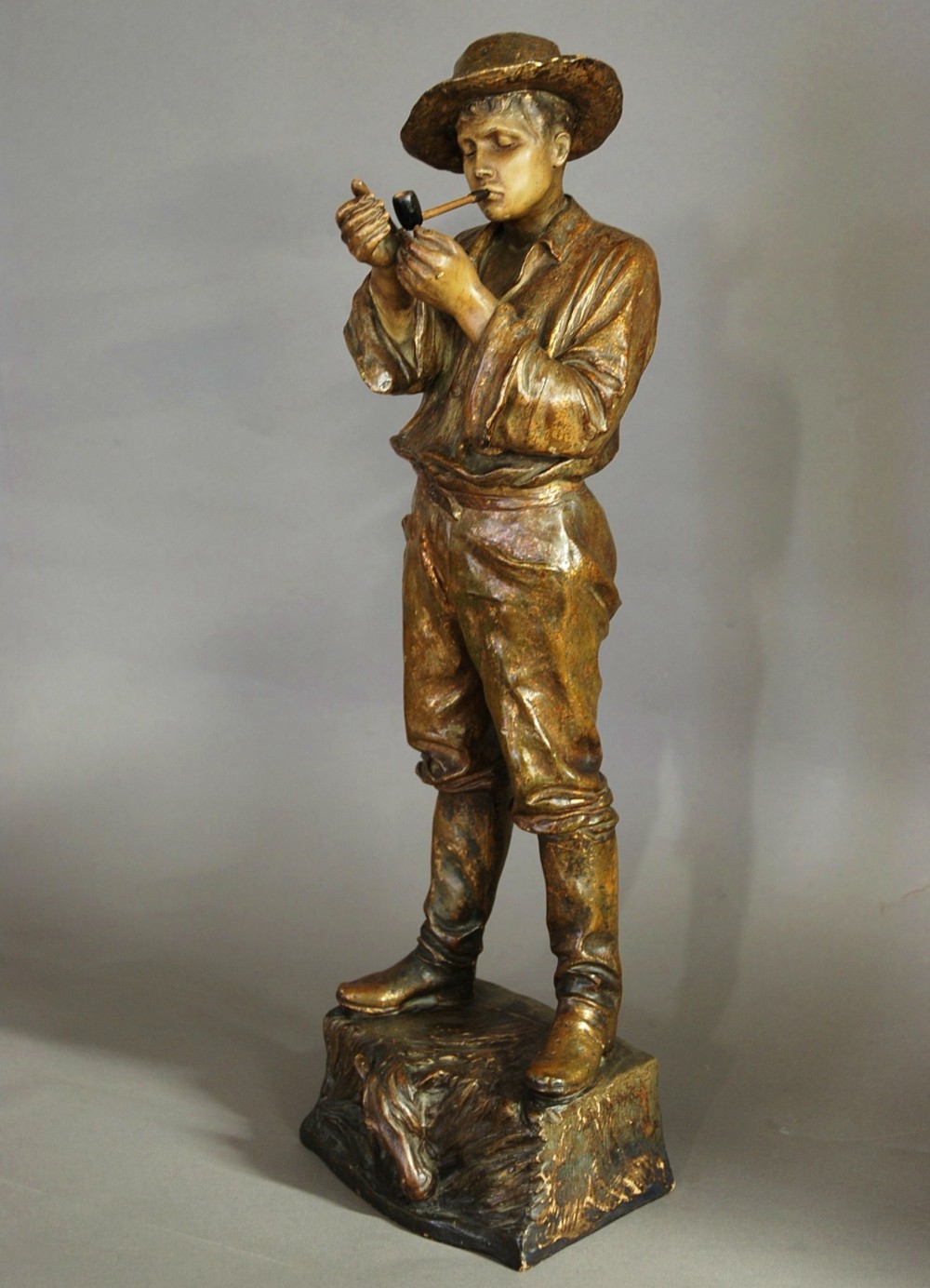 A bronzed terracotta figure of a standing man wearing a hat and lighting a pipe on a naturalistic base.