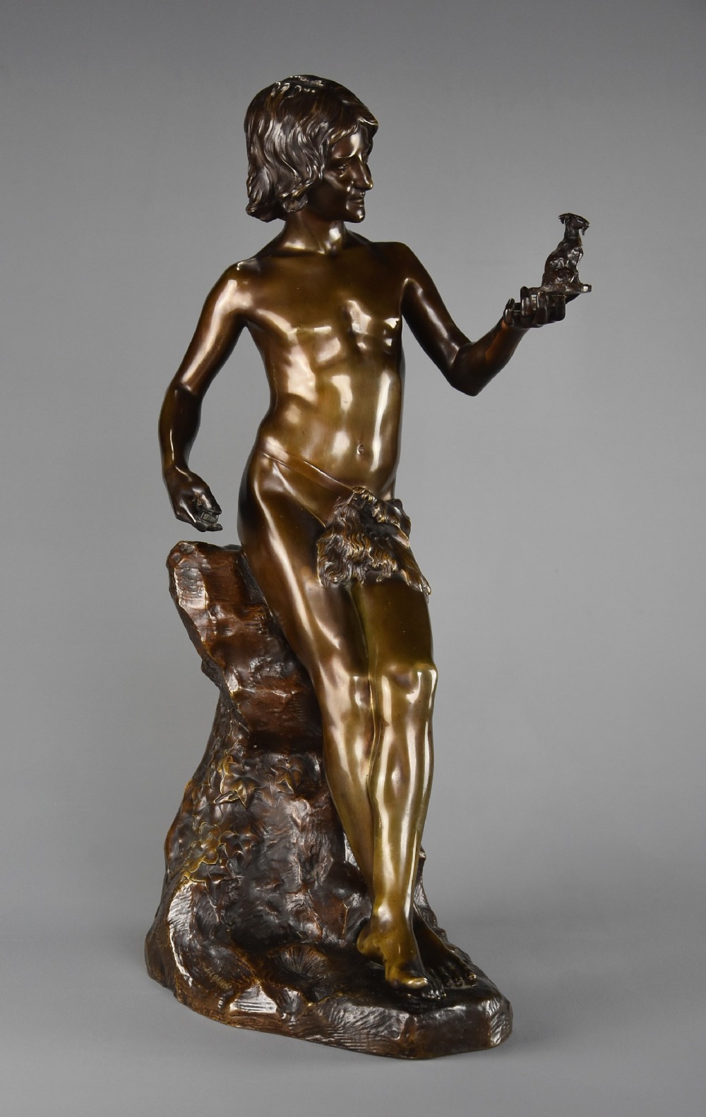 A dark brown patinated bronze sculpture depicting a standing youth leaning against a rocky base while holding a small figurine of a goat in his raised left hand.