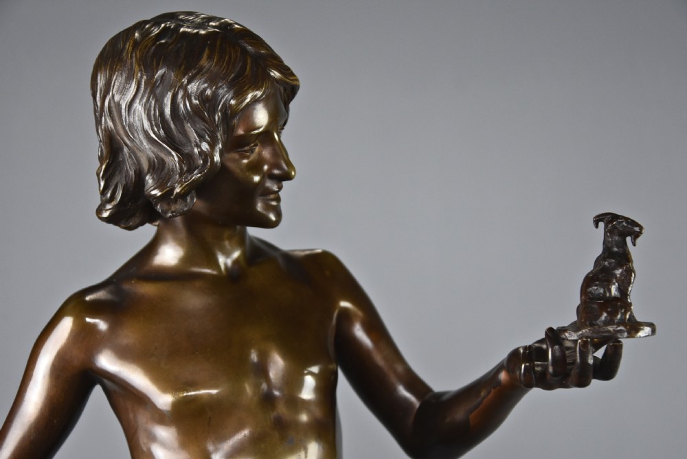 A dark brown patinated bronze sculpture depicting a standing youth leaning against a rocky base while holding a small figurine of a goat in his raised left hand. View XI