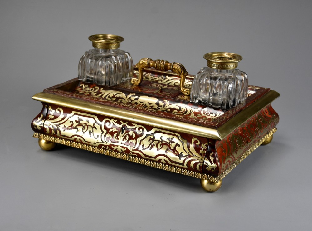 A mid-19th-century desk stand crafted with red tortoiseshell and brass scrollwork inlay, featuring two glass inkwells with brass lids, a central handle, and brass ball feet.