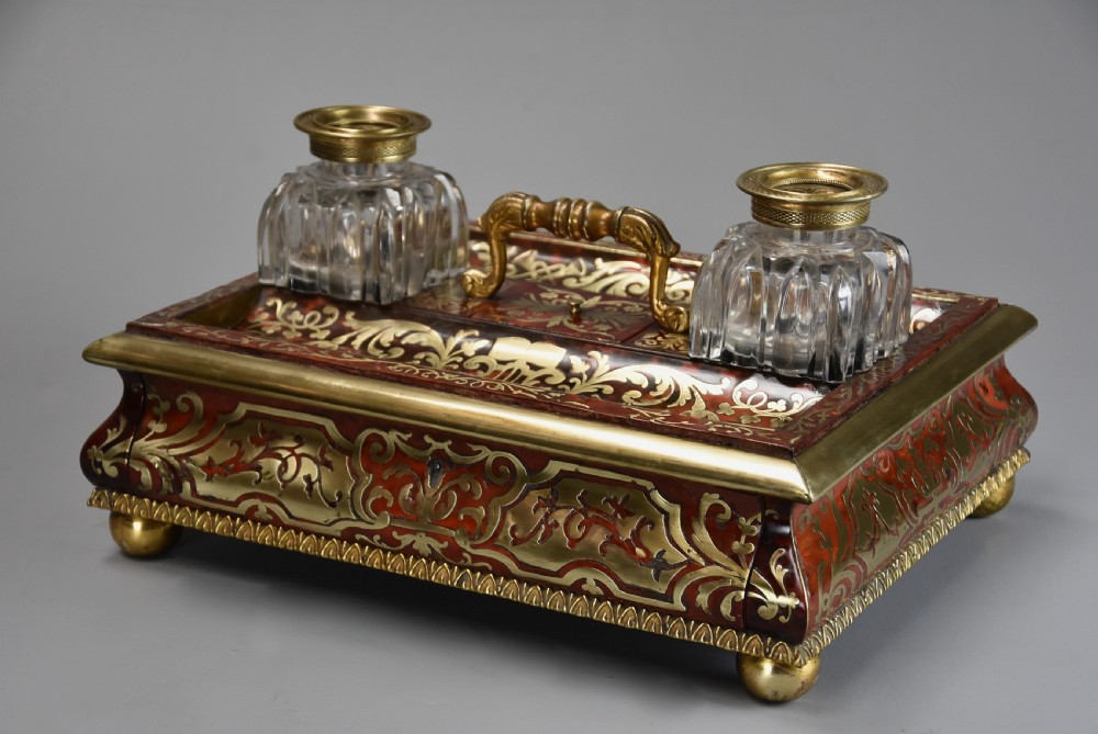 A mid-19th-century desk stand crafted with red tortoiseshell and brass scrollwork inlay, featuring two glass inkwells with brass lids, a central handle, and brass ball feet. View II