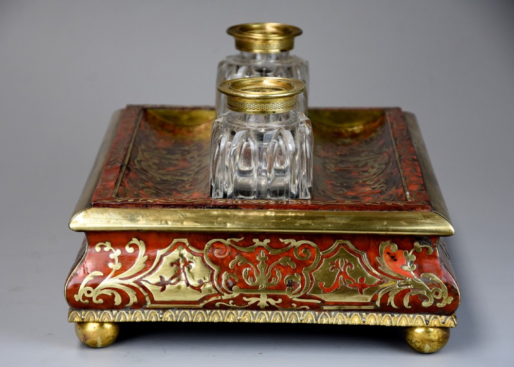 A mid-19th-century desk stand crafted with red tortoiseshell and brass scrollwork inlay, featuring two glass inkwells with brass lids, a central handle, and brass ball feet. View VI