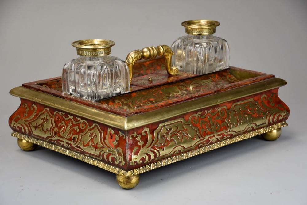 A mid-19th-century desk stand crafted with red tortoiseshell and brass scrollwork inlay, featuring two glass inkwells with brass lids, a central handle, and brass ball feet. View IX