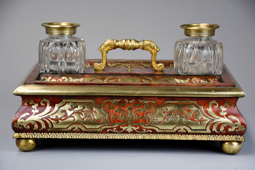 A mid-19th-century desk stand crafted with red tortoiseshell and brass scrollwork inlay, featuring two glass inkwells with brass lids, a central handle, and brass ball feet. View X
