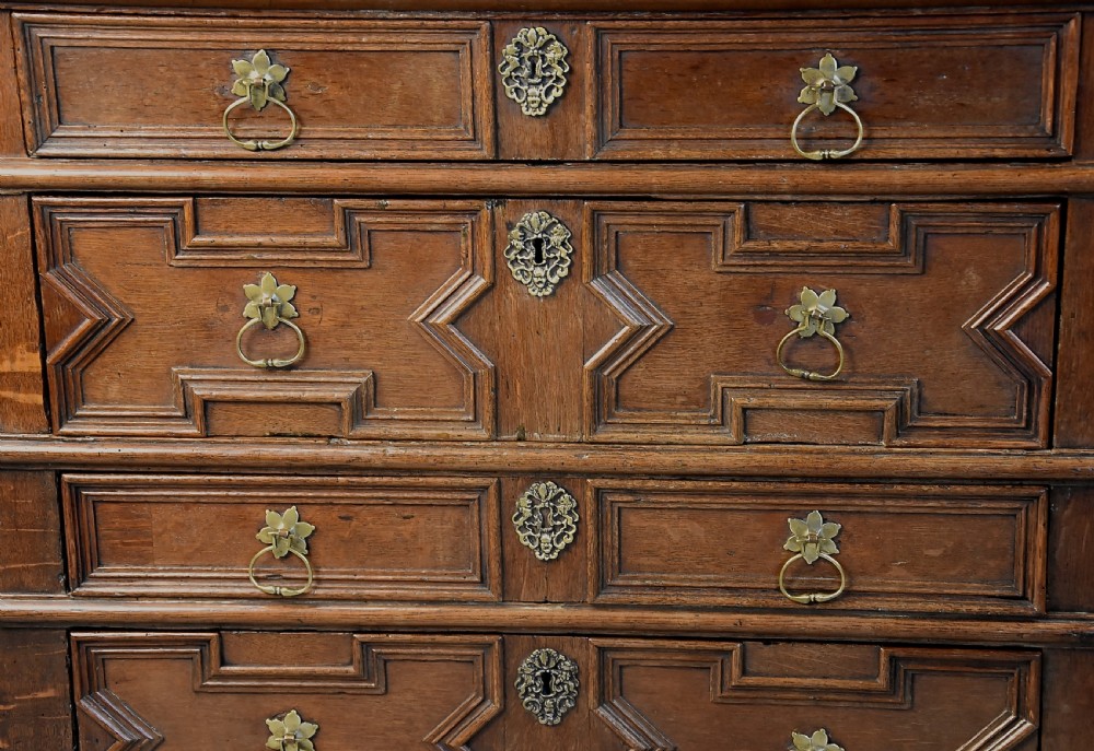 A late 17th-century oak chest of drawers featuring geometric moulded drawer fronts, brass drop handles, and decorative brass escutcheons. View VII