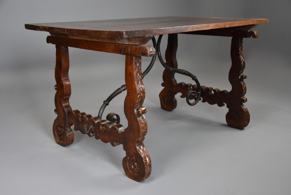 A solid walnut trestle table featuring carved decorative legs, hand-forged wrought iron stretchers, and a dark polished top.