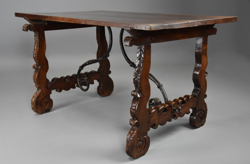 A solid walnut trestle table featuring carved decorative legs, hand-forged wrought iron stretchers, and a dark polished top. View V