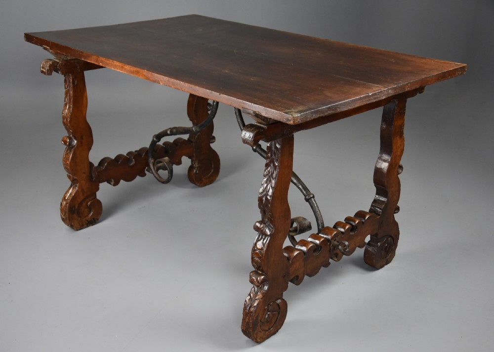 A solid walnut trestle table featuring carved decorative legs, hand-forged wrought iron stretchers, and a dark polished top. View VI