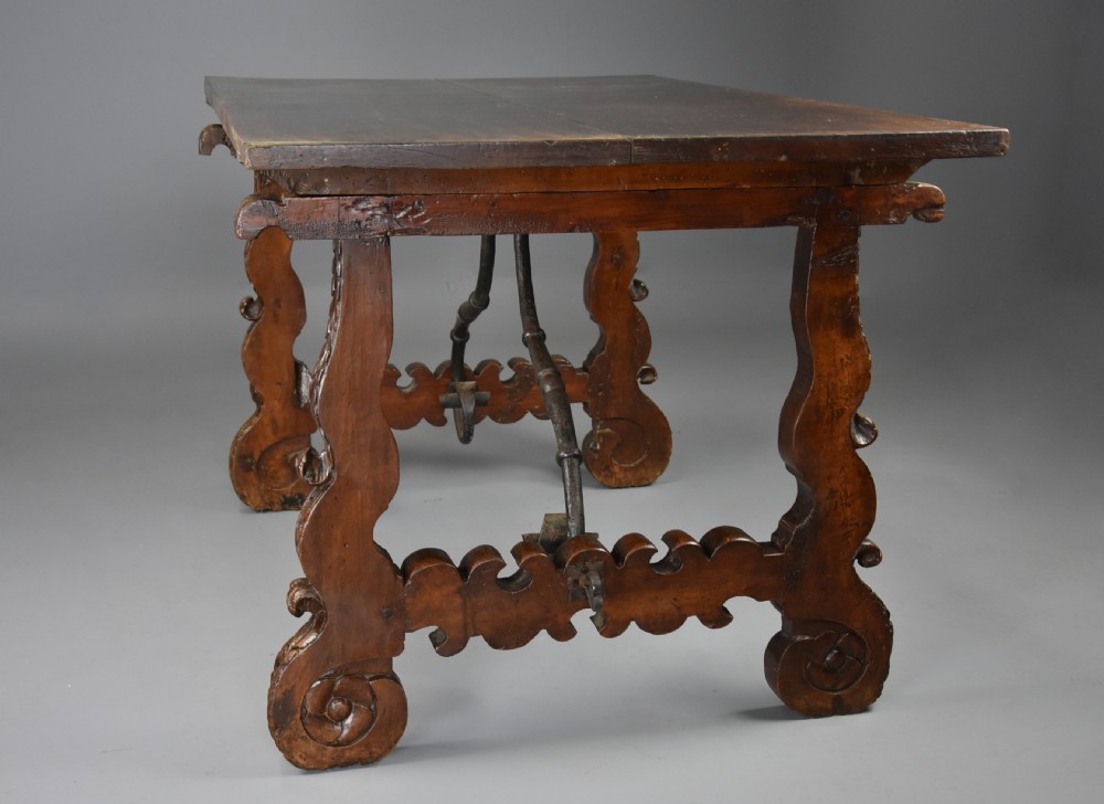 A solid walnut trestle table featuring carved decorative legs, hand-forged wrought iron stretchers, and a dark polished top. View IX