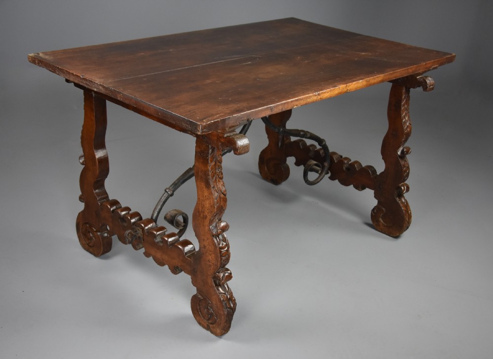 A solid walnut trestle table featuring carved decorative legs, hand-forged wrought iron stretchers, and a dark polished top. View X