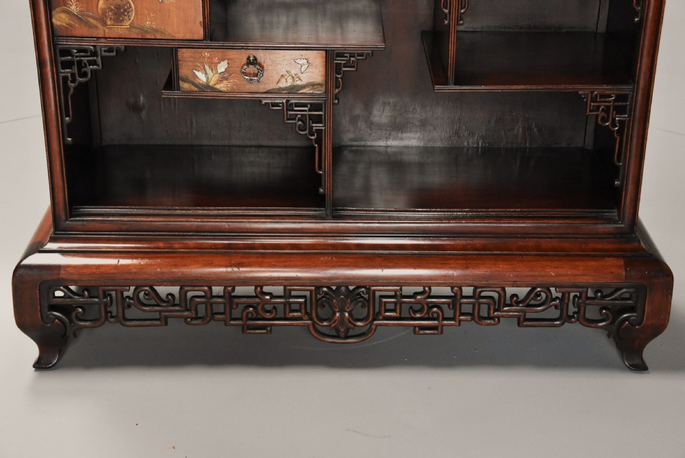 A late 19th-century dark padouk display cabinet featuring asymmetrical shelving, reticulated fretwork, and inlaid pictorial panel doors. View XI