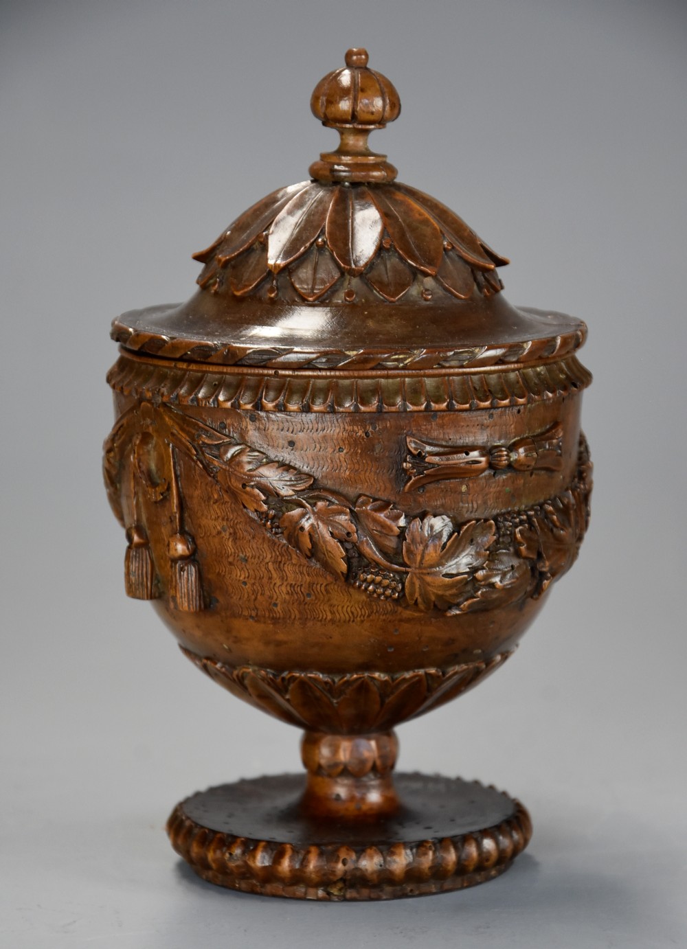 A late eighteenth-century carved walnut lidded urn featuring floral garlands, tassel motifs, a leaf-carved dome lid with finial, resting on a turned circular base.