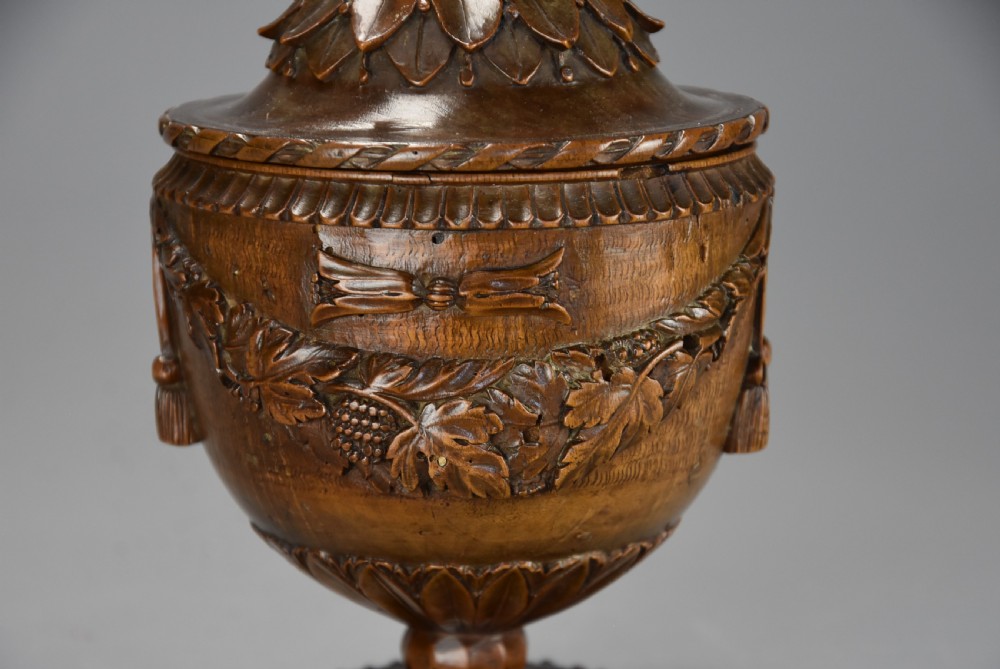 A late eighteenth-century carved walnut lidded urn featuring floral garlands, tassel motifs, a leaf-carved dome lid with finial, resting on a turned circular base. View V