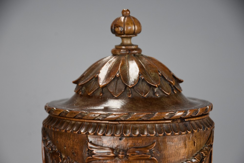 A late eighteenth-century carved walnut lidded urn featuring floral garlands, tassel motifs, a leaf-carved dome lid with finial, resting on a turned circular base. View VI