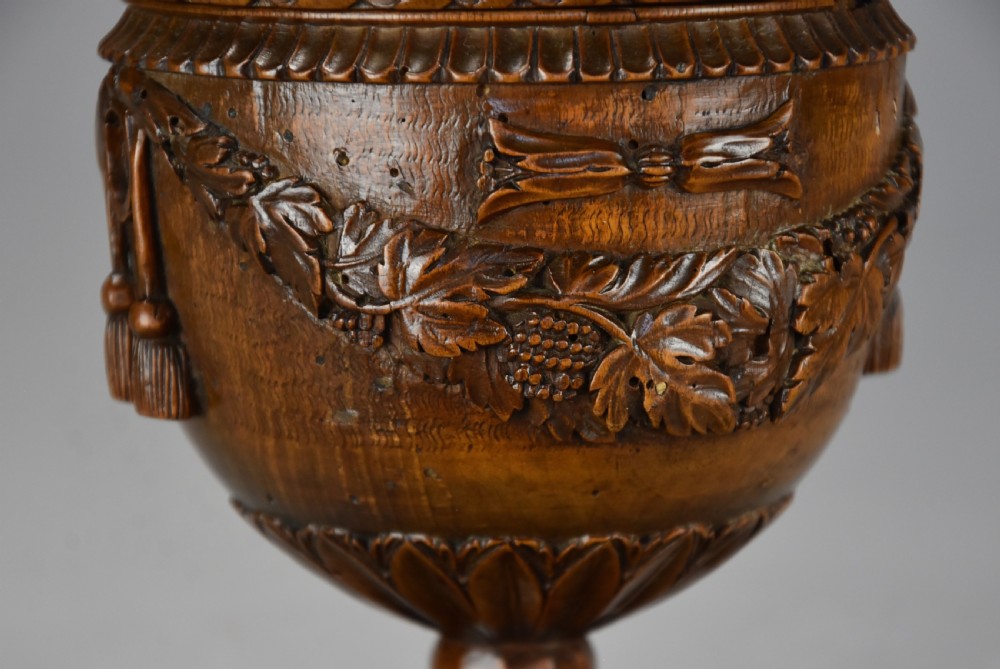 A late eighteenth-century carved walnut lidded urn featuring floral garlands, tassel motifs, a leaf-carved dome lid with finial, resting on a turned circular base. View VII