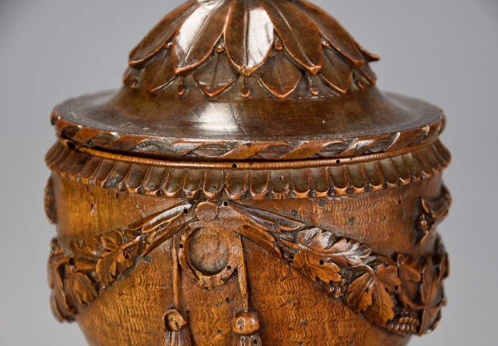 A late eighteenth-century carved walnut lidded urn featuring floral garlands, tassel motifs, a leaf-carved dome lid with finial, resting on a turned circular base. View XI