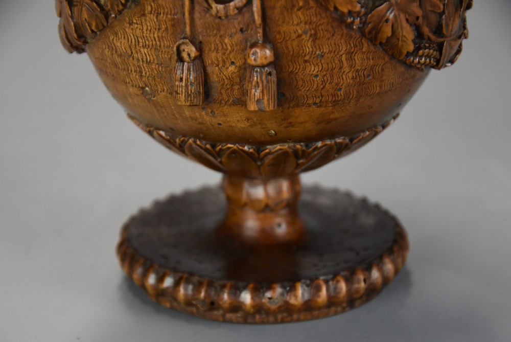 A late eighteenth-century carved walnut lidded urn featuring floral garlands, tassel motifs, a leaf-carved dome lid with finial, resting on a turned circular base. View XII