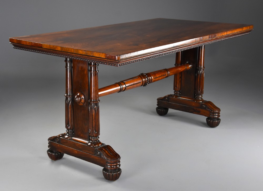A William IV rosewood centre writing table featuring a rectangular top, beaded border, carved end supports, a central turned stretcher, and lobed feet.