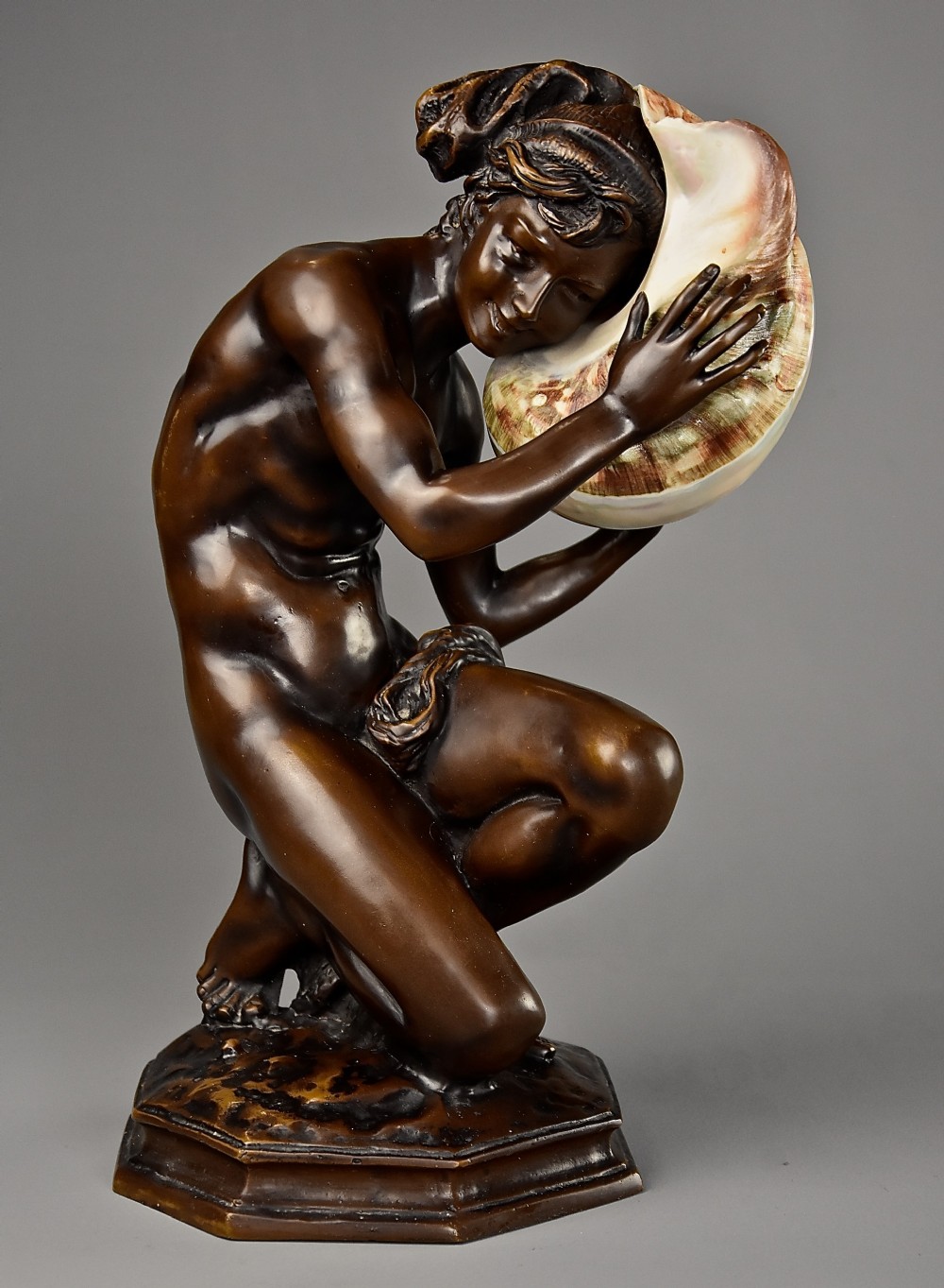 A patinated dark bronze sculpture depicting a youth kneeling and holding a large seashell to his ear on an octagonal base.