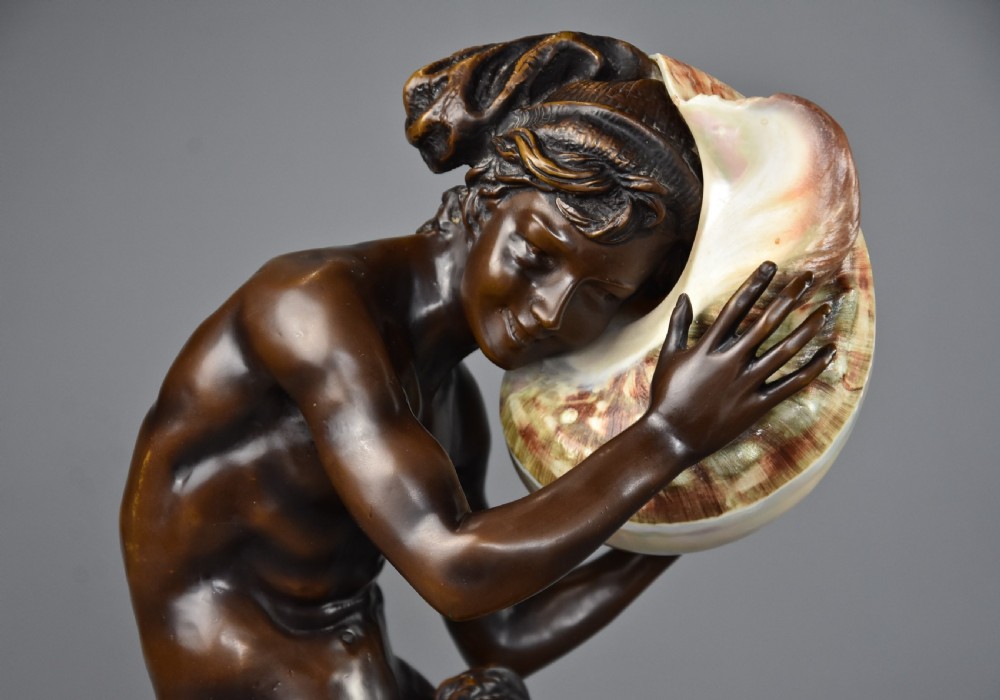 A patinated dark bronze sculpture depicting a youth kneeling and holding a large seashell to his ear on an octagonal base. View IV