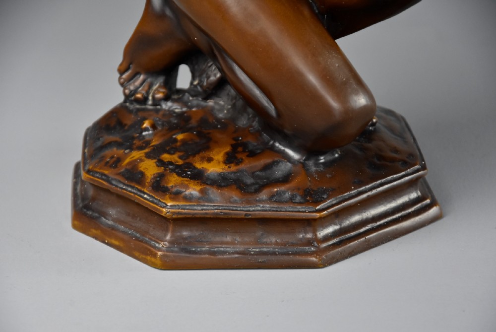 A patinated dark bronze sculpture depicting a youth kneeling and holding a large seashell to his ear on an octagonal base. View XI