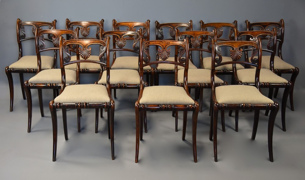 A set of thirteen early nineteenth-century Regency dining chairs featuring carved anthemion backrests, curved legs, and light neutral upholstered seat cushions.