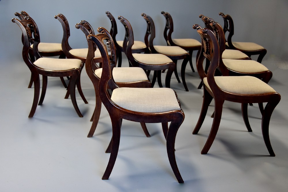 A set of thirteen early nineteenth-century Regency dining chairs featuring carved anthemion backrests, curved legs, and light neutral upholstered seat cushions. View VI