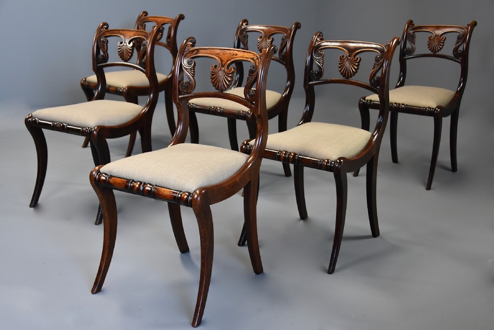 A set of thirteen early nineteenth-century Regency dining chairs featuring carved anthemion backrests, curved legs, and light neutral upholstered seat cushions. View X