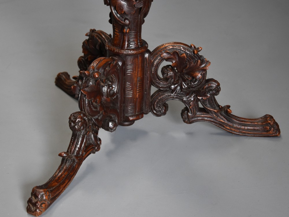 A circular late 19th-century dark brown linden wood centre table featuring intricate foliate carvings on the top, a decorated pedestal, and a tripod base. View IX