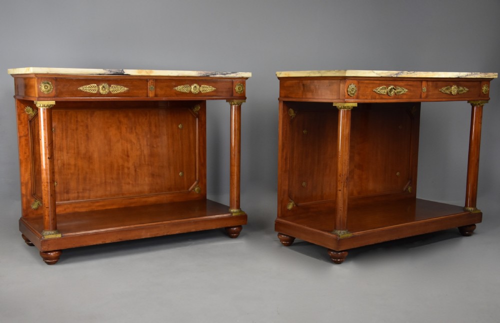 A pair of 19th-century French Empire style mahogany console tables featuring light marble tops, gilt bronze mounts, and cylindrical front columns.
