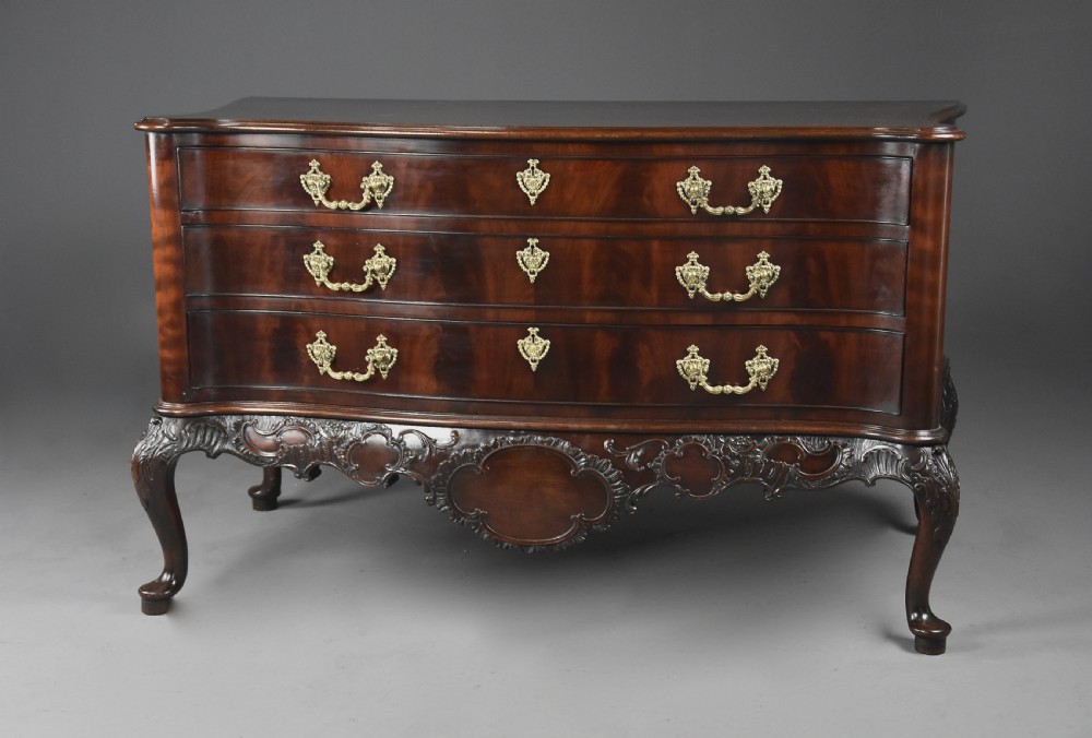 A late nineteenth-century dark flame mahogany serpentine chest of drawers featuring three graduated drawers with decorative brass handles and a carved lower apron. View II
