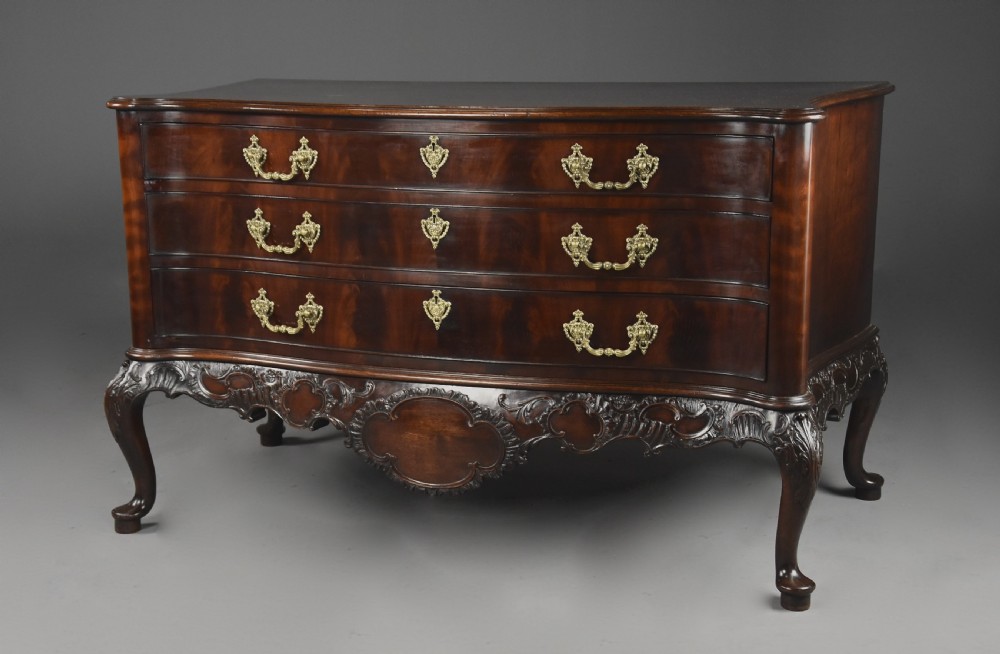 A late nineteenth-century dark flame mahogany serpentine chest of drawers featuring three graduated drawers with decorative brass handles and a carved lower apron. View III