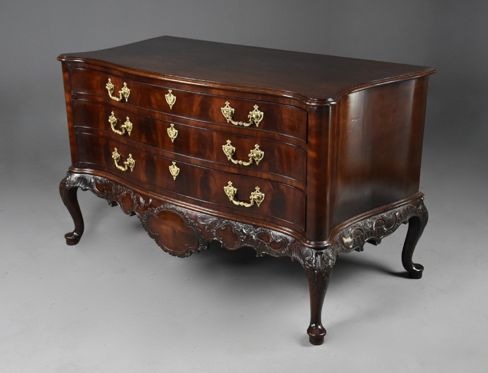A late nineteenth-century dark flame mahogany serpentine chest of drawers featuring three graduated drawers with decorative brass handles and a carved lower apron. View IV