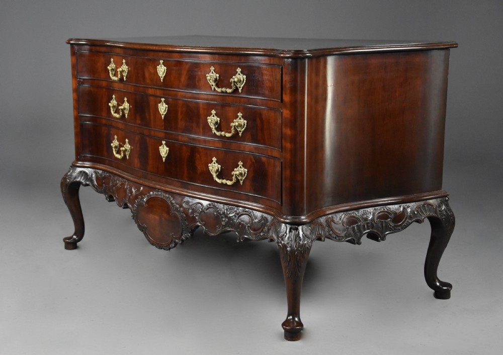 A late nineteenth-century dark flame mahogany serpentine chest of drawers featuring three graduated drawers with decorative brass handles and a carved lower apron. View V