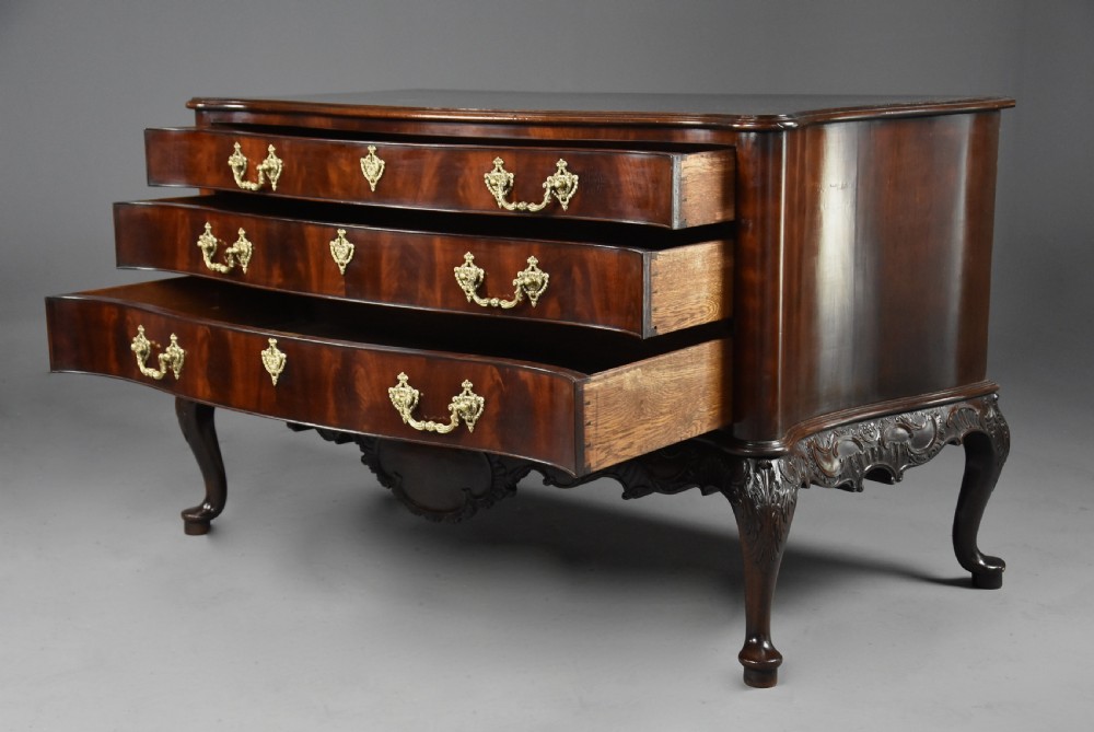 A late nineteenth-century dark flame mahogany serpentine chest of drawers featuring three graduated drawers with decorative brass handles and a carved lower apron. View IX