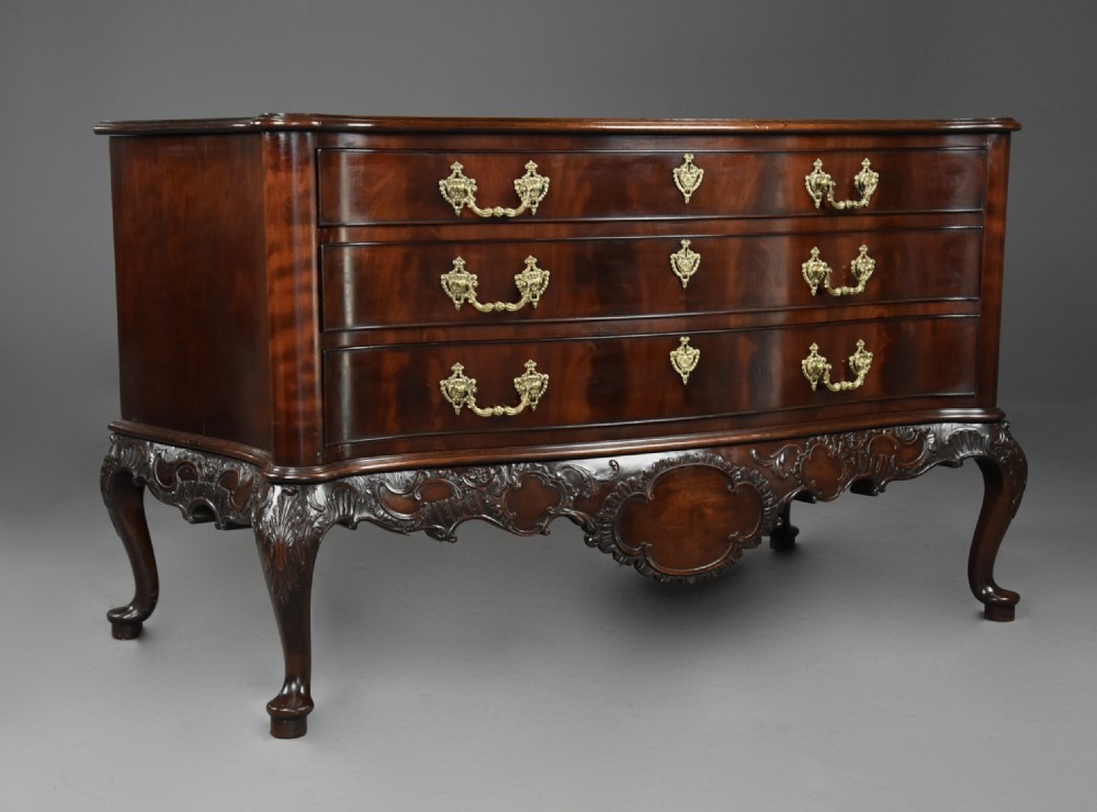 A late nineteenth-century dark flame mahogany serpentine chest of drawers featuring three graduated drawers with decorative brass handles and a carved lower apron. View X