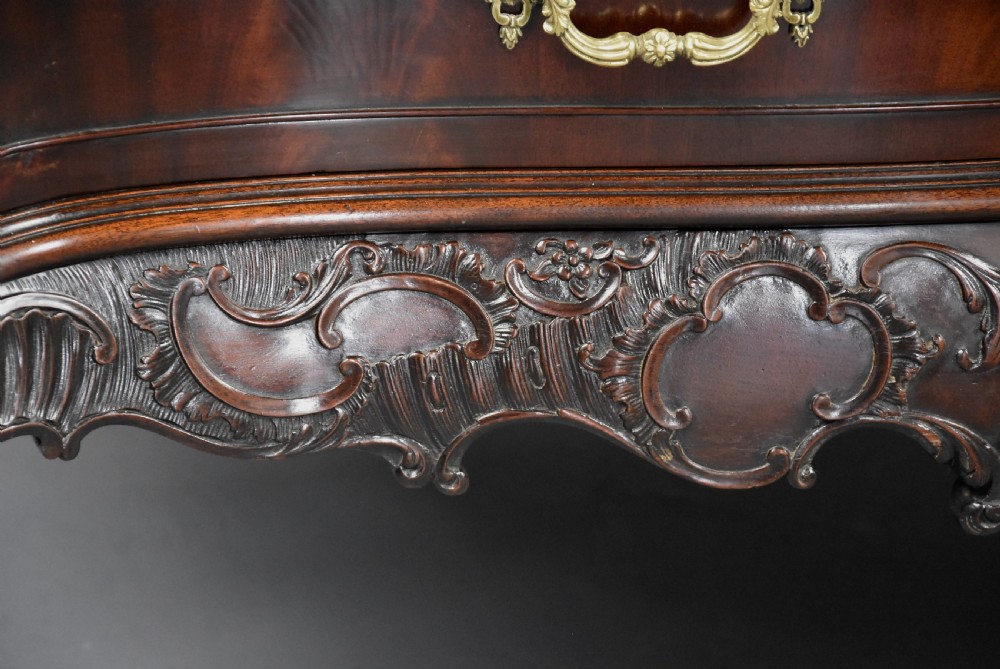 A late nineteenth-century dark flame mahogany serpentine chest of drawers featuring three graduated drawers with decorative brass handles and a carved lower apron. View XII