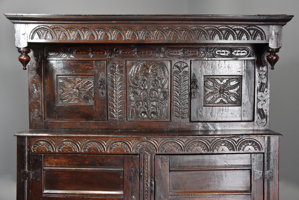 A 17th-century dark oak press cupboard featuring upper carved panel doors, overhanging canopy with pendant drops, and lower paneled double doors. View IV