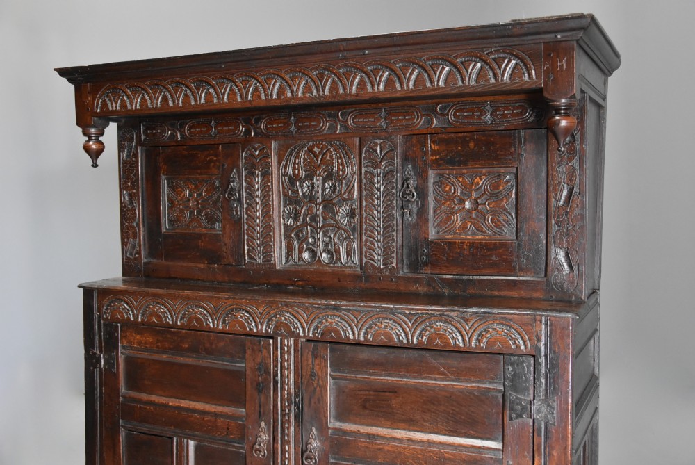A 17th-century dark oak press cupboard featuring upper carved panel doors, overhanging canopy with pendant drops, and lower paneled double doors. View V