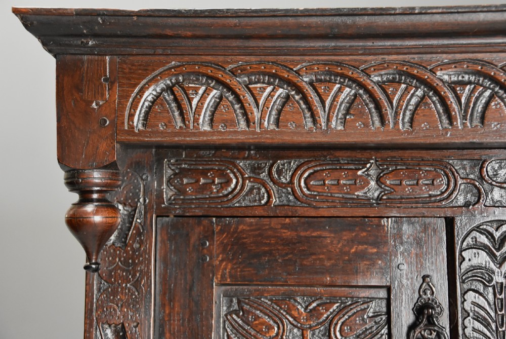 A 17th-century dark oak press cupboard featuring upper carved panel doors, overhanging canopy with pendant drops, and lower paneled double doors. View VI